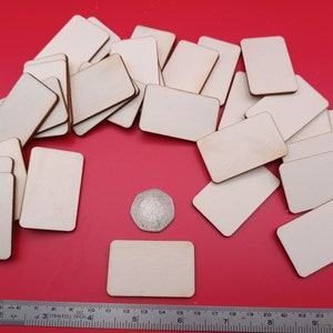 Może przedstawiać: Stosik 25 nieoszlifowanych, prostokątnych drewnianych półfabrykatów, o wymiarach około 5 cm x 7,5 cm, na czerwonym tle. Dla porównania rozmiarów dołączono brytyjską monetę.