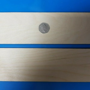 May include: Two rectangular pieces of unfinished wood, one slightly larger than the other. The larger piece has a coin on top of it for scale.