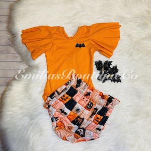May include: An orange Halloween-themed baby outfit set. The set includes a ribbed bodysuit with flutter sleeves and a bat applique, and matching patterned bloomers. A black glitter bat hair clip is also included.