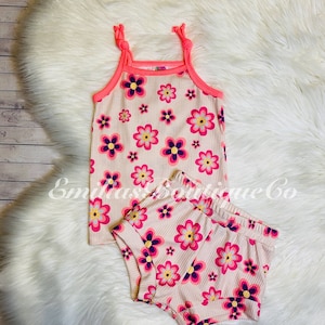 May include: A two-piece baby outfit with a pink tank top and matching shorts. Both pieces feature a floral print with pink, yellow, and purple flowers on a cream background. The tank top has pink straps that tie at the shoulders.