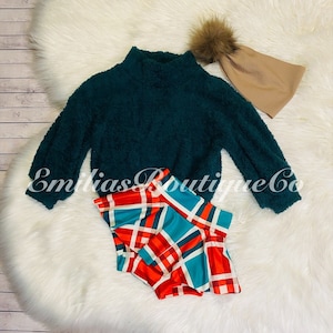 May include: A teal fuzzy sweater with long sleeves and a mock neck. The sweater is paired with a red, white, and blue plaid skirt. A brown beanie with a fluffy pom pom is also included.