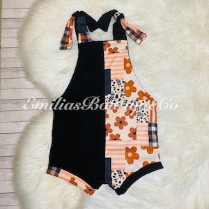 May include: Black and orange patchwork baby romper with a floral print and plaid straps. The romper has a knotted detail at the straps.