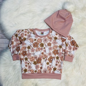 May include: A pink long-sleeved sweatshirt with a print of cookies, milk bottles, and flowers. It comes with a matching pink beanie with a white pom-pom.