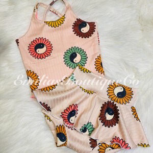 May include: A pink and white two-piece outfit with a sunflower and yin-yang design. The top is a tank top with criss-cross straps and the bottom is a ruffled skirt.