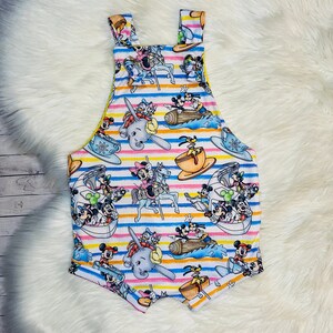 May include: White and yellow striped overalls with a colorful print of various Disney characters, including Mickey Mouse, Minnie Mouse, Donald Duck, Goofy, and Pluto. The characters are depicted in various scenes, such as riding a carousel, sailing on a boat, and riding an elephant.