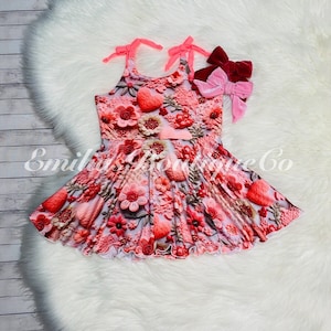 May include: A pink and red floral dress with a heart and flower embellishments. The dress has tie straps and a ruffled hem. Two velvet bows in pink and burgundy are attached to the dress. The dress is on a white fluffy surface.