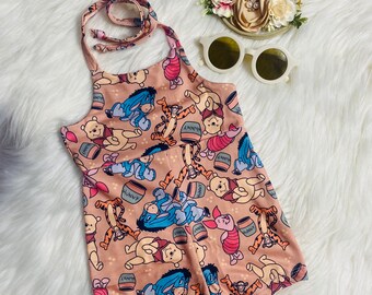 May include: A Winnie the Pooh themed halter top romper with a brown background and a variety of characters including Winnie the Pooh, Tigger, Piglet, and Eeyore. The romper has a halter top style with a tie at the neck.