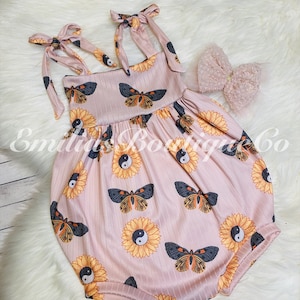 May include: A pink baby onesie with a sunflower and butterfly print. The onesie has adjustable straps that tie at the shoulders. The onesie has a yin yang symbol in the center of each sunflower.