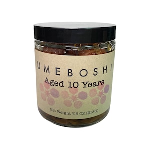 May include: A clear glass jar with a black lid, filled with a preserved food product. The label reads "UMEBOSHI Aged 10 Years" with a floral design. The net weight is 7.5 OZ (213g).