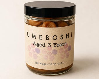 Op de afbeelding: Een doorzichtige glazen pot met een zwarte deksel gevuld met bruine ingelegde pruimen. Het beige etiket bevat de tekst UMEBOSHI Aged 3 Years, drie lichtpaarse en roze bloemillustraties en het nettogewicht van 213g op de voorzijde gedrukt.