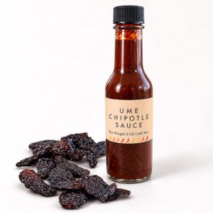 May include: A clear glass bottle of Ume Chipotle Sauce with a black cap and a tan label reading Ume Chipotle Sauce Net Weight 5 OZ (148 ML) with a row of chili icons. A pile of dark, dried chipotle peppers sits next to the bottle.