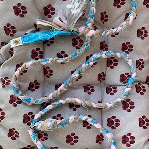 May include: A braided dog toy made from different fabrics, including blue, pink, and brown. The toy is in the shape of a circle and is lying on a gray and red patterned surface.