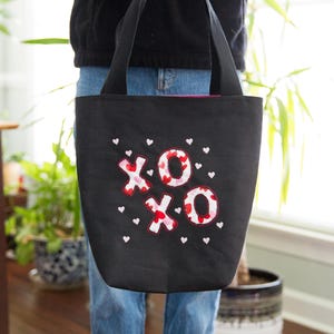 May include: Black tote bag with embroidered "XOXO" design in red, white, and pink. The bag features small white heart accents. The tote has black handles and is held by a person wearing blue jeans.