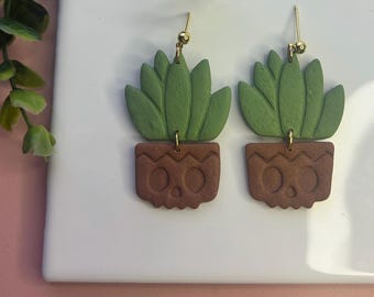 Puede incluir: Pendientes hechos a mano con un diseño de suculenta verde en una maceta marrón. Los pendientes tienen ganchos dorados y se exhiben sobre una superficie blanca con un fondo rosa. La suculenta tiene múltiples hojas verdes.