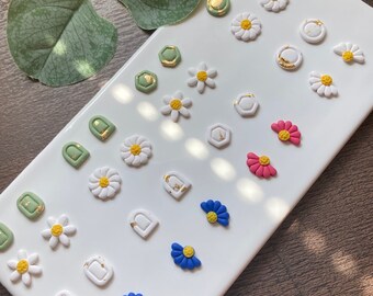 Puede incluir: Una colección de pendientes de arcilla polimérica coloridos en varias formas, incluyendo flores, arcos y diseños geométricos. Los pendientes presentan tonos blancos, verdes, rosas y azules con acentos de pan de oro. Están dispuestos sobre una bandeja rectangular blanca.