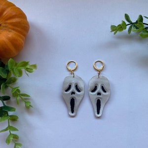 Könnte beinhalten: Ein Paar weiße Ohrhänger mit einem schwarzen Scream-Masken-Design. Die Ohrringe haben goldene Ringe.