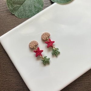 Puede incluir: Un par de pendientes colgantes con una concha, una estrella de mar roja y un dije de tortuga verde. Los pendientes están hechos de arcilla polimérica y tienen un gancho dorado.