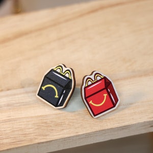 Op de afbeelding: Twee houten pinnen in de vorm van McDonald's-dozen. Eén is rood met een gele glimlach en de andere is zwart met een gele glimlach.