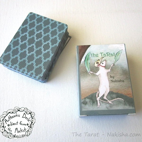 May include: A deck of tarot cards with a blue and white geometric pattern on the back. The box features a cartoon rat wearing a crown and holding two green leaves. The text on the box reads "the TaRat by Nakisha".