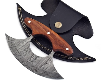 May include: A Damascus steel knife with a curved blade and a wooden handle, resting on a black leather sheath. The knife features a unique pattern on the blade and is designed for cutting. The sheath has a decorative button.