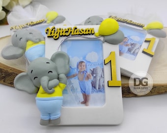May include: A white picture frame with a gray elephant figurine holding a yellow balloon. The frame has a photo of a baby boy in a blue outfit. The frame also has the number "1" and the name "Yiğit Hasan" written in gold letters.