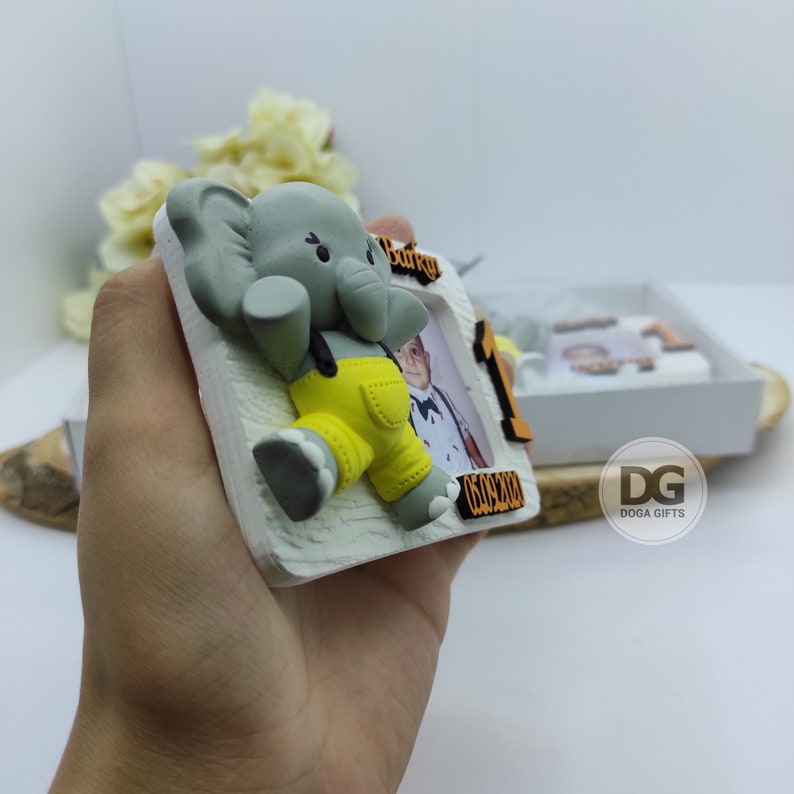 May include: A white photo frame with a gray elephant figurine wearing yellow overalls. The frame has a space for a photo and the number "1" with the date "05.09.2020" printed on it. The frame is held in a hand.