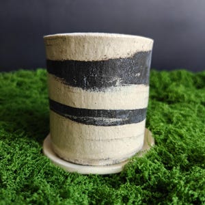 Puede incluir: Una maceta cilíndrica de cerámica con un acabado mate en color blanco roto y dos bandas horizontales de pinceladas negras pintadas a mano que rodean el exterior. La maceta se asienta sobre un plato circular a juego con una superficie lisa.