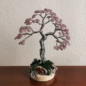 May include: A wire sculpture of a tree with pink leaves and a twisted trunk. The tree is sitting on a wooden base with moss and a large, red stone.
