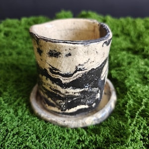 May include: A small cylindrical ceramic planter with a matching circular saucer. The exterior features a marbled pattern of contrasting black and off-white clay, creating a textured, organic appearance with an uneven, hand-formed rim.