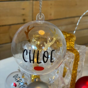 May include: Clear glass Christmas bauble with a reindeer silhouette and the name "CHLOE" written in black lettering. The bauble has a red nose and gold antlers.