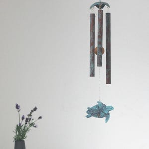 May include: A decorative wind chime with a turtle design. The chime features three copper-colored tubes with a verdigris finish, suspended from a blue cord. A turquoise-colored turtle hangs below.