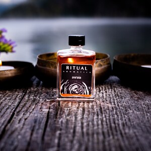 May include: A small glass bottle of amber-colored liquid with a black label that reads "RITUAL AROMATICS asraia". The bottle is sitting on a wooden surface with two candles in front of it.