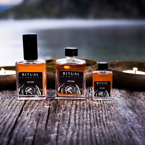 May include: Three glass bottles of Ritual Aromatics essential oil in various sizes. The bottles are amber colored and have black labels with white text. The text on the labels reads "Ritual Aromatics" and "Astraea". The bottles are sitting on a wooden surface with two candles in the background.