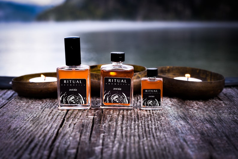 May include: Three glass bottles of Ritual Aromatics essential oil in various sizes. The bottles are amber colored and have black labels with white text. The text on the labels reads "Ritual Aromatics" and "Astraea". The bottles are sitting on a wooden surface with two candles in the background.