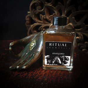 Peut inclure: Un petit flacon en verre de liquide ambré avec un bouchon noir. Le flacon porte une étiquette noire avec du texte blanc qui dit "RITUAL AROMATICS" et "MORGANA".