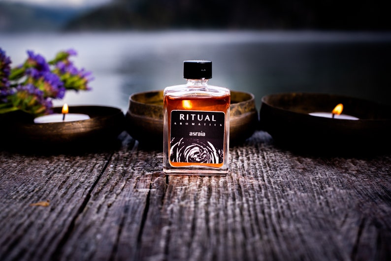 May include: A small glass bottle of amber-colored liquid with a black label that reads "RITUAL AROMATICS asraia". The bottle is sitting on a wooden surface with two candles in front of it.
