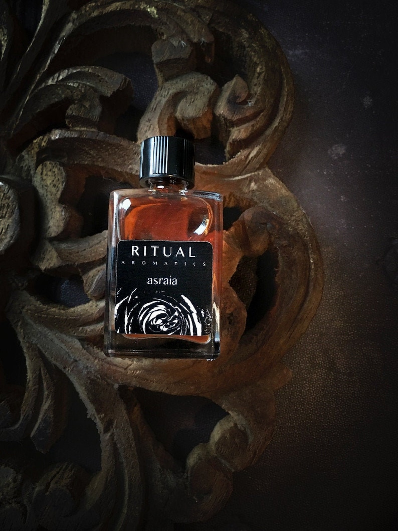 May include: A small, clear glass bottle with a black cap and label. The label reads "RITUAL AROMATILS asraia". The bottle is sitting on a dark brown wooden surface with intricate carvings.