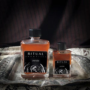 May include: Two glass bottles of amber-colored liquid with black caps and labels. The labels read "Ritual Aromatics" and "Asraia". The bottles are on a silver tray with a lace tablecloth underneath.