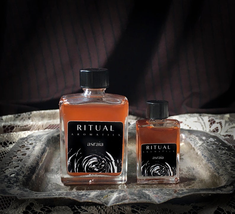 May include: Two glass bottles of amber-colored liquid with black caps and labels. The labels read "Ritual Aromatics" and "Asraia". The bottles are on a silver tray with a lace tablecloth underneath.