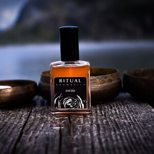 May include: A brown glass bottle of Ritual Aromatics asraia perfume with a black cap, sitting on a wooden surface with two lit candles in brass bowls.