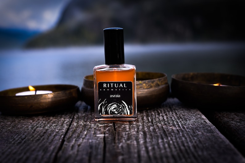 May include: A brown glass bottle of Ritual Aromatics asraia perfume with a black cap, sitting on a wooden surface with two lit candles in brass bowls.