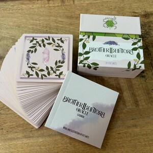 May include: A deck of Brother II Brother Grace Oracle cards with a pink and green floral design. The cards are fanned out and the box is closed. The box is white with green and pink floral designs and the text "Brother II Brother Grace Oracle" in black.