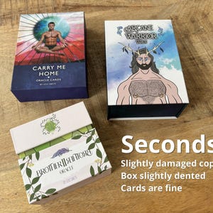 May include: Three oracle card decks on a wooden surface. The decks include "Carry Me Home," "Arcane Warrior Tarot," and "Brotherhood Oracle." The image also includes the text "Seconds," indicating slightly damaged copies.