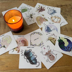 May include: A set of 12 oracle cards with illustrations of various plants, animals, and crystals. The cards are white with black text and are laid out on a wooden surface. A lit candle is in the foreground.
