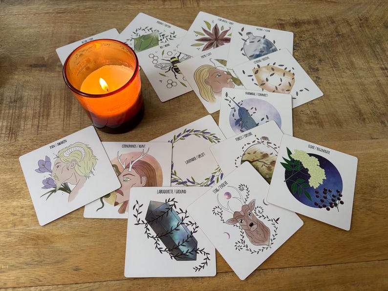 May include: A set of 12 oracle cards with illustrations of various plants, animals, and crystals. The cards are white with black text and are laid out on a wooden surface. A lit candle is in the foreground.