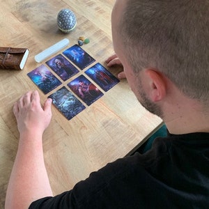 May include: A person is reading a tarot card spread on a wooden table. The cards are arranged in a grid pattern and feature colorful illustrations. The person is wearing a black shirt and has short brown hair.