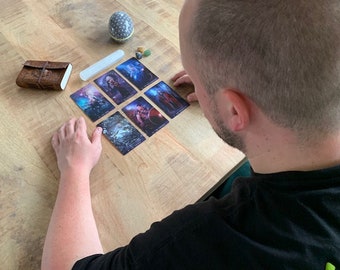 May include: A person is reading a tarot card spread on a wooden table. The cards are arranged in a grid pattern and feature colorful illustrations. The person is wearing a black shirt and has short brown hair.