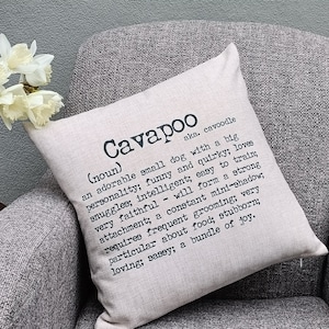 May include: A beige throw pillow with the word "Cavapoo" printed in black, along with a definition of the word. The definition reads: "(noun) aka. cavoodle an adorable small dog with a big personality; funny and quirky; loves snuggles; intelligent; easy to train; very faithful - will form a strong attachment; a constant mini-shadow; requires frequent grooming; very particular about food; stubborn; loving; sassy; a bundle of joy."