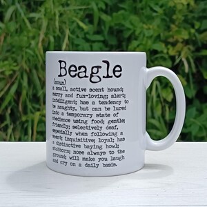 May include: White ceramic mug featuring the word "Beagle" in black, accompanied by a definition of the breed. The mug has a curved handle and is set against a blurred green backdrop.