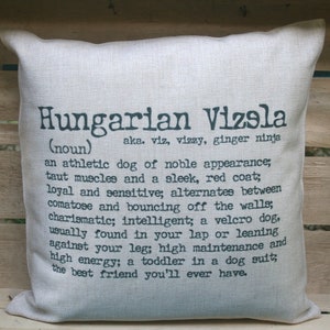 Może przedstawiać: Kwadratowa, kremowa poduszka z napisem "Hungarian Vizsla" w ciemnoszarym kolorze i definicją. Definicja opisuje cechy psa, w tym jego atletyczną budowę i energię.